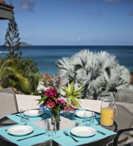An inviting outdoor dining table set with vibrant blue placemats, fresh flowers, and a pitcher of orange juice, overlooking a stunning ocean view.
