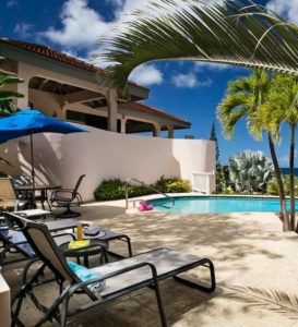 Relaxing poolside lounge area with a blue umbrella, lounge chairs, and palm trees providing shade at Adagio Villa Resort.