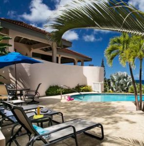 Relaxing poolside lounge area with a blue umbrella, lounge chairs, and palm trees providing shade at Adagio Villa Resort.