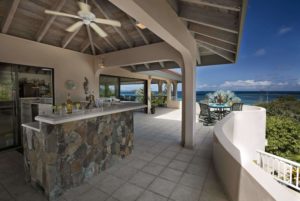 A stunning outdoor bar area featuring a stone countertop, ceiling fans, and large windows offering breathtaking ocean views.