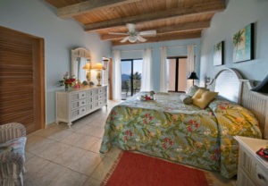 A serene tropical master bedroom featuring a king-sized bed with floral bedding, large windows offering ocean views, and warm wooden accents.