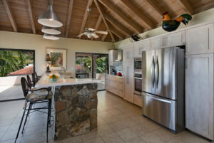A stylish tropical kitchen featuring white cabinetry, a stone island bar, stainless steel appliances, and large windows offering stunning ocean views.