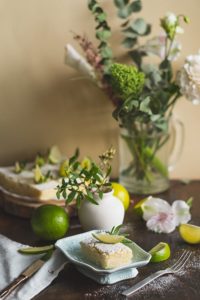 A slice of lime cake served on a plate, surrounded by fresh limes, floral decorations, and a vase of greenery.