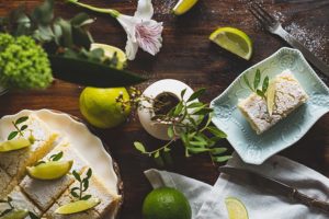 A slice of lime cake dusted with powdered sugar, garnished with fresh lime slices and mint leaves, served on a decorative plate surrounded by limes and floral accents.