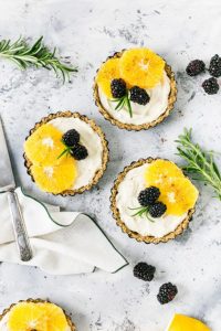 Three mini tarts filled with creamy lemon curd, topped with blackberries, orange slices, and fresh rosemary sprigs.