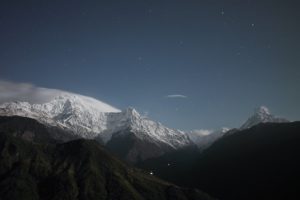 A breathtaking view of snow-capped mountains under a starry night sky, with wispy clouds adding depth to the scene.