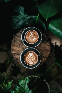 Two cups of latte with intricate leaf and heart designs in the foam, served on a wooden tray surrounded by lush green leaves.