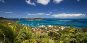 A breathtaking panoramic view of a tropical bay, featuring turquoise waters dotted with sailboats, lush green hills, and vibrant red-roofed houses.