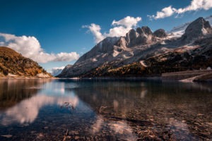 A serene lake with crystal-clear waters reflecting towering mountains and vibrant autumn trees under a bright blue sky.