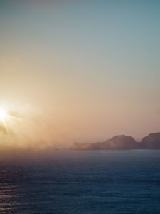 A serene sunset scene over the ocean, with misty cliffs and distant islands silhouetted against the warm hues of the sky.
