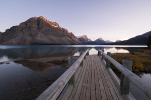 A serene wooden dock extending into a tranquil lake, reflecting the majestic mountains and warm sunset hues.