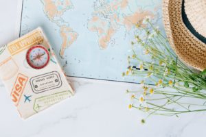 Travel essentials including a passport cover, compass, and world map, surrounded by flowers and a straw hat.