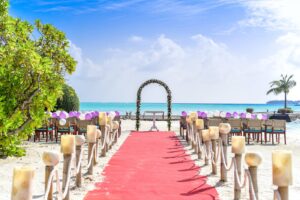 Romantic beach wedding setup with a red carpet, candlelit aisle, and floral arch overlooking the ocean.