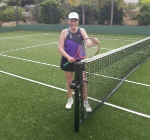 Smiling woman holding a tennis racket on the Mahoe Bay community tennis court in Virgin Gorda, British Virgin Islands, standing near the net on a lush green artificial turf court.